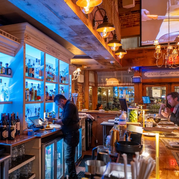 A bartender works behind a bar with blue-lit shelves of liquor, while patrons sit at the counter.
