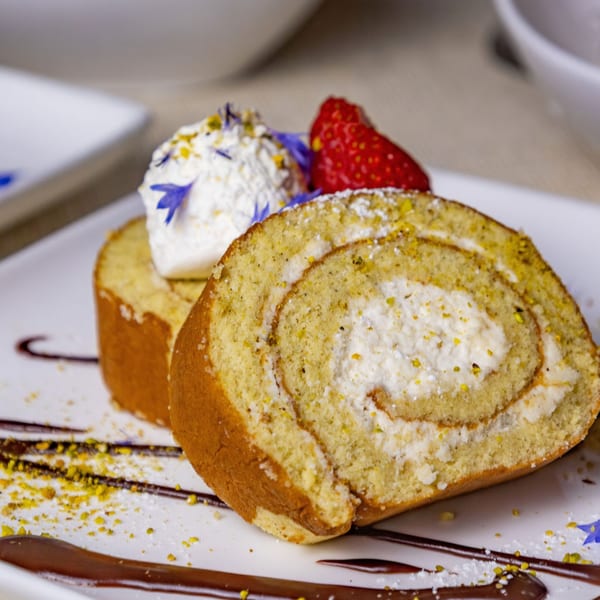 Two slices of pistachio roll cake with whipped cream, strawberry, and blue edible flowers.