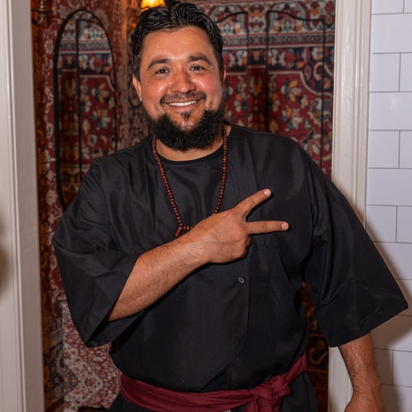 Smiling Darvish Kitchen chef in black attire and red sash, making a peace sign.