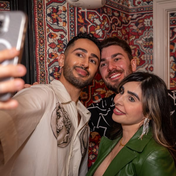 Three smiling people taking a selfie in a room with a patterned Middle Eastern rug on the wall.
