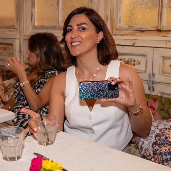 A smiling woman in a white top holds up a phone with a heart-patterned case at Darvish Kitchen.