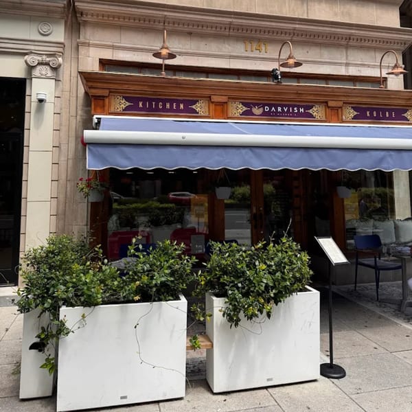 The exterior of Darvish Kitchen, a Middle Eastern restaurant, with a blue awning and large planters.