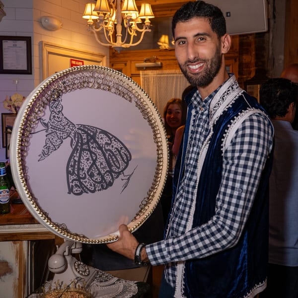 Smiling man in a blue velvet vest holding a round tray with a calligraphic design.