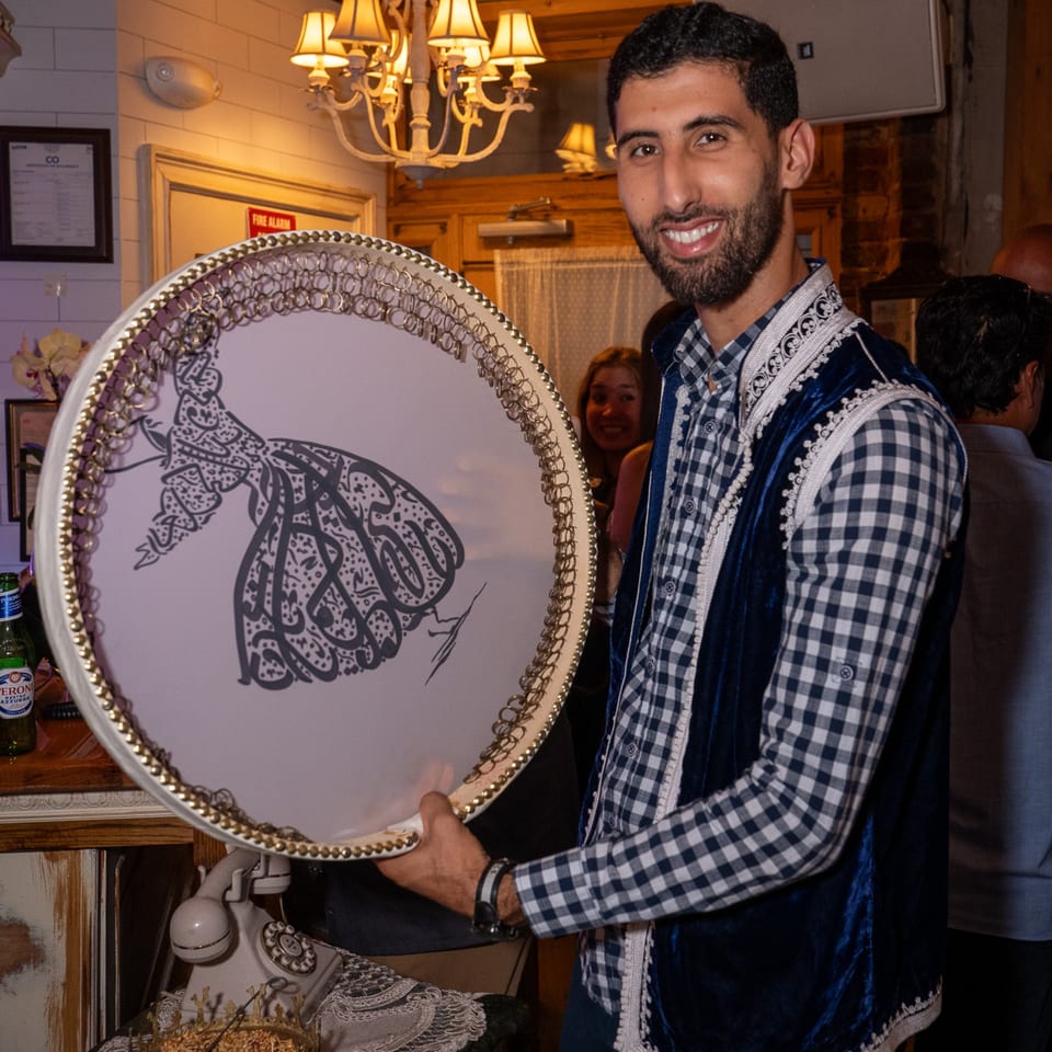 Smiling man in a blue velvet vest holding a round tray with a calligraphic design.