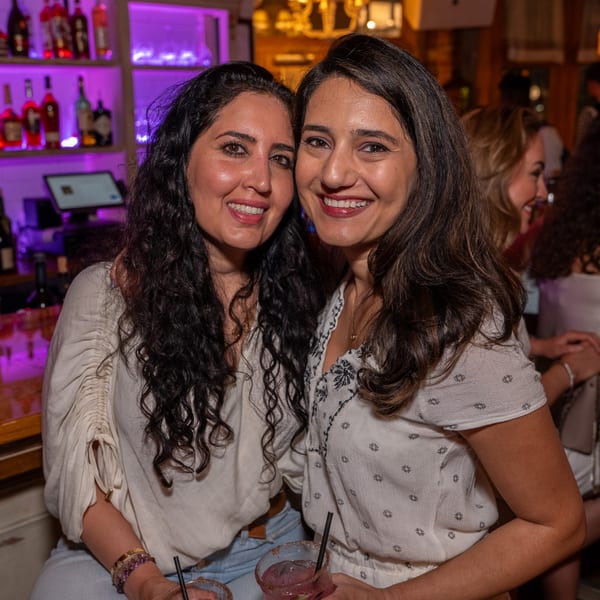 Two smiling women with drinks at a bar with purple lighting, enjoying the Darvish Kitchen atmosphere.
