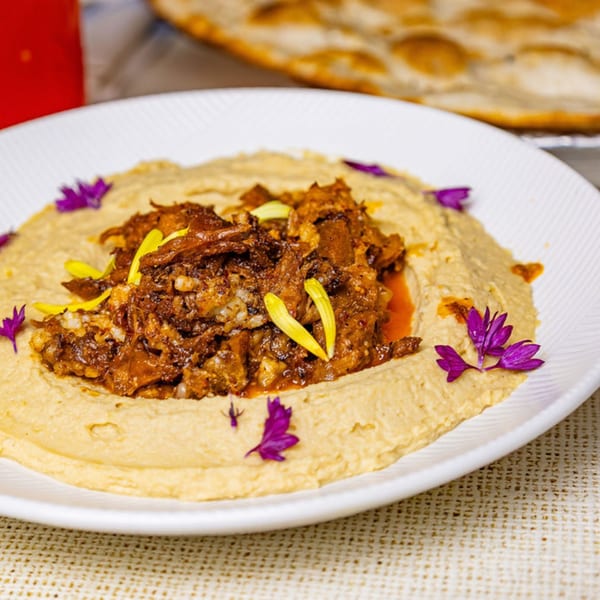 Hummus topped with shredded meat, garnished with purple and yellow edible flowers, on a white plate.