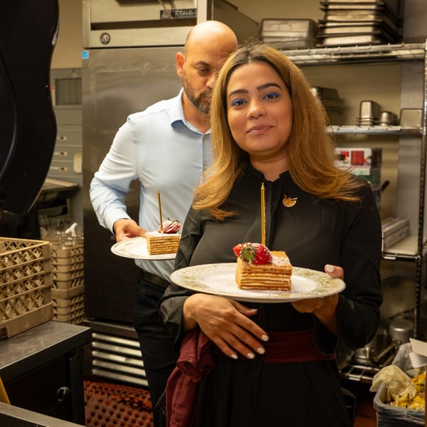 Two Darvish Kitchen staff members in a kitchen holding plates of layered cake with candles and strawberries.