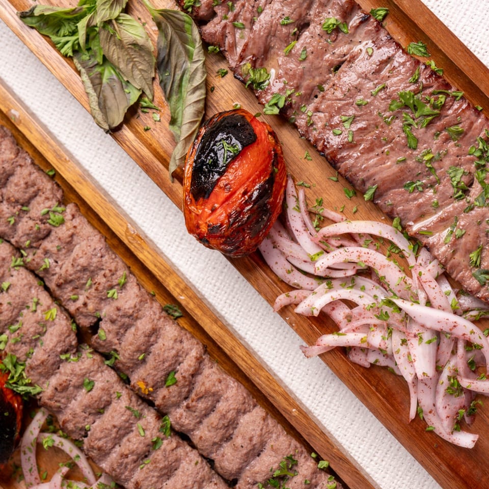 Two wooden trays of Darvish Kitchen's grilled meats, charred tomato, sliced onions, and fresh herbs.