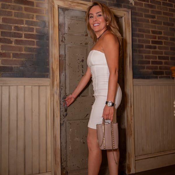 A smiling woman in a white dress opens a rustic wooden door, holding a studded pink handbag.