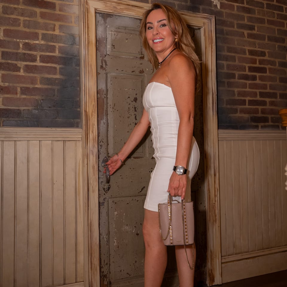 A smiling woman in a white dress opens a rustic wooden door, holding a studded pink handbag.