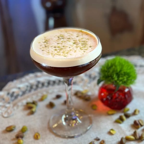 A Darvish Kitchen cocktail with foam and green sprinkles, surrounded by pistachios and a red vase.