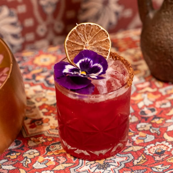 A vibrant red cocktail garnished with a dried orange slice and purple pansy on a patterned rug.