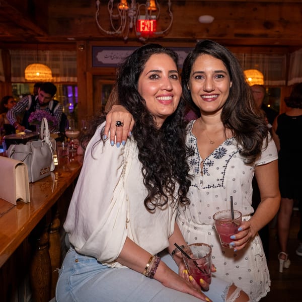 Two smiling women holding cocktails pose at a bar inside Darvish Kitchen, with an exit sign visible.
