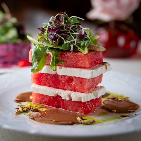 Stacked watermelon and feta salad with microgreens, drizzled with sauce and pistachios on a white plate.