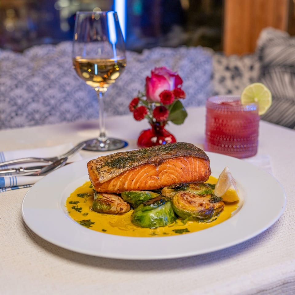 Grilled salmon with roasted brussels sprouts in a yellow sauce, white wine, and a pink cocktail.
