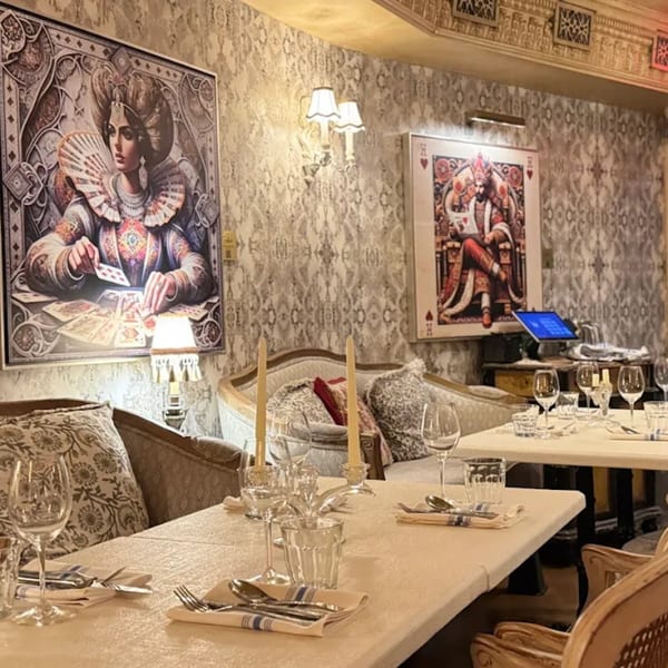 Elegant dining room with set tables, patterned wallpaper, and large framed artwork of playing card figures.