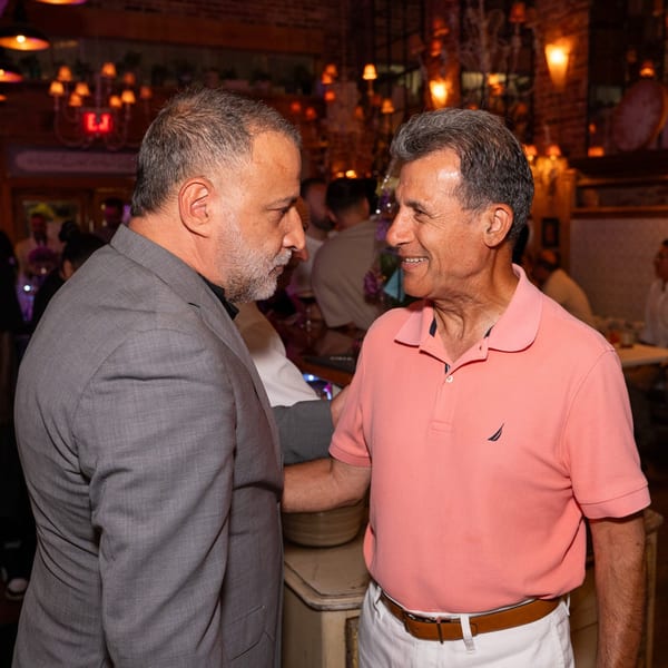 Two middle-aged men, one in a grey suit and one in a pink polo, talking in a dimly lit restaurant.