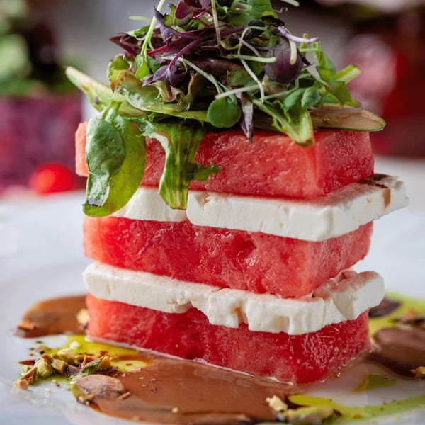 Stacked watermelon and feta cheese salad, topped with greens and drizzled with sauce and pistachios.
