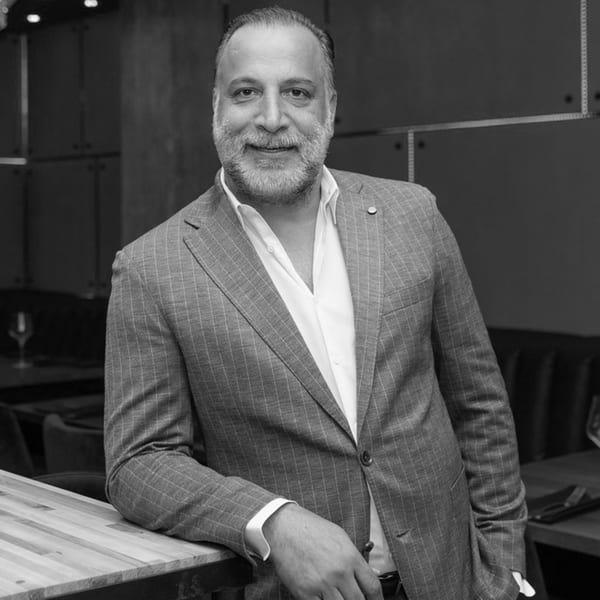 Smiling man with a beard in a pinstriped suit jacket, leaning on a wooden counter.