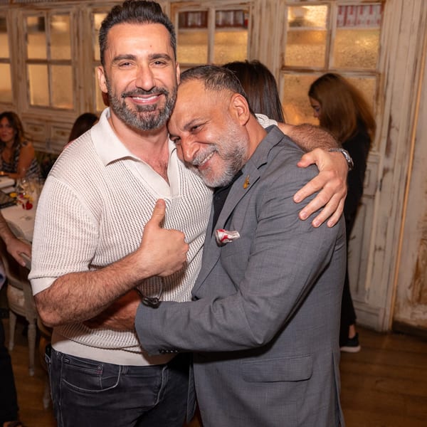 Two smiling men embrace warmly, one giving a thumbs-up, in a restaurant setting.