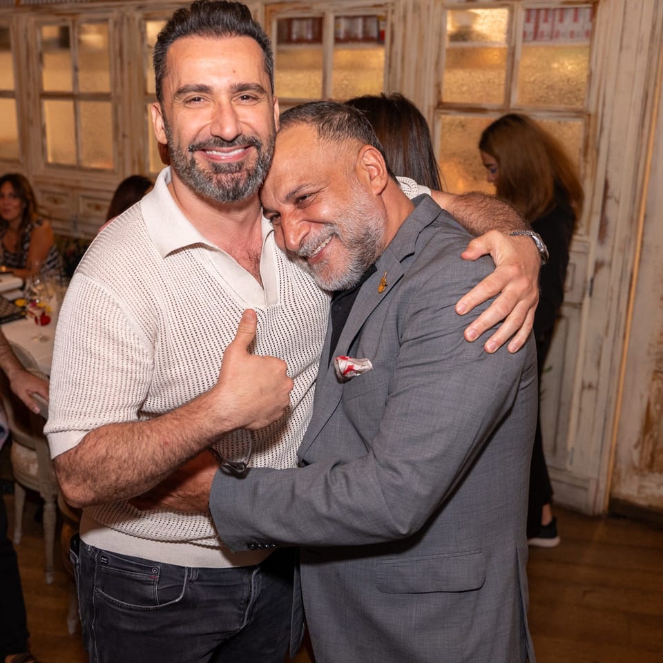 Two smiling men embrace warmly, one giving a thumbs-up, in a restaurant setting.