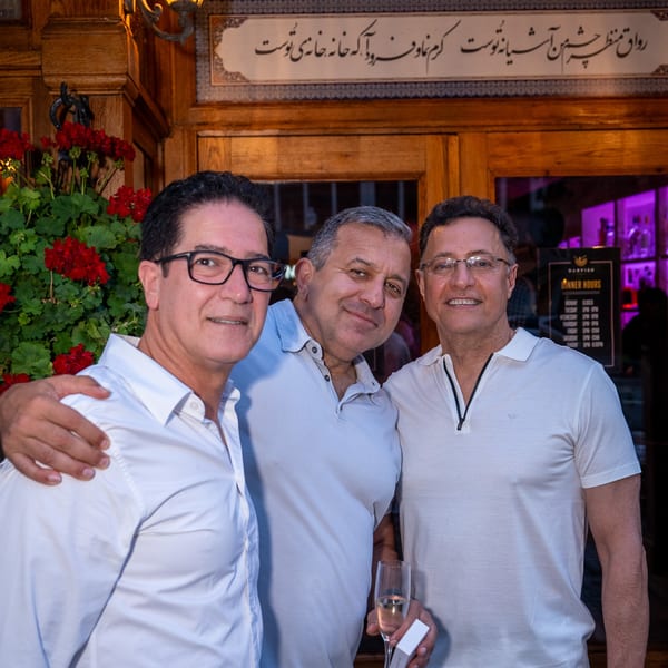 Three men smiling, one holding a champagne flute, in front of Darvish Kitchen's ornate wooden entrance.