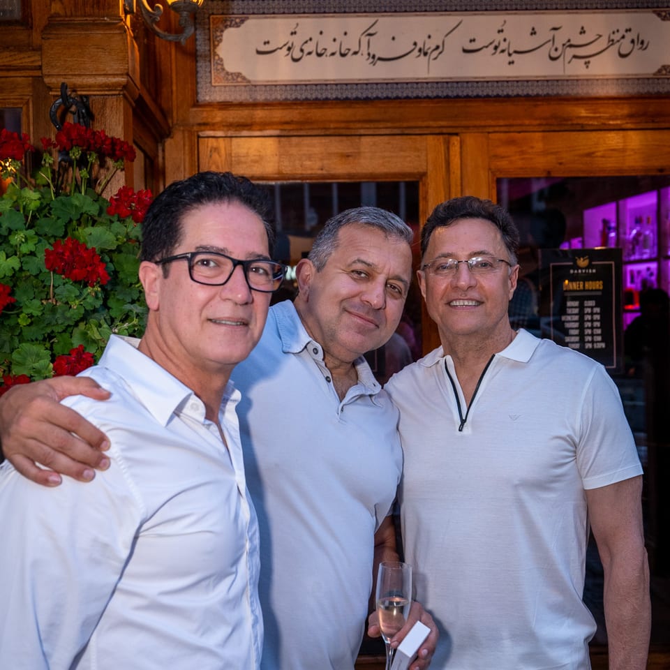 Three men smiling, one holding a champagne flute, in front of Darvish Kitchen's ornate wooden entrance.