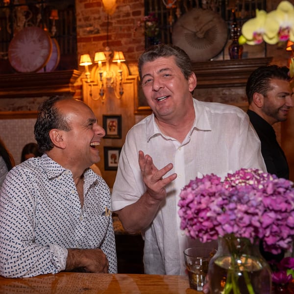 Two men laughing and talking at a bar, with purple flowers in the foreground.