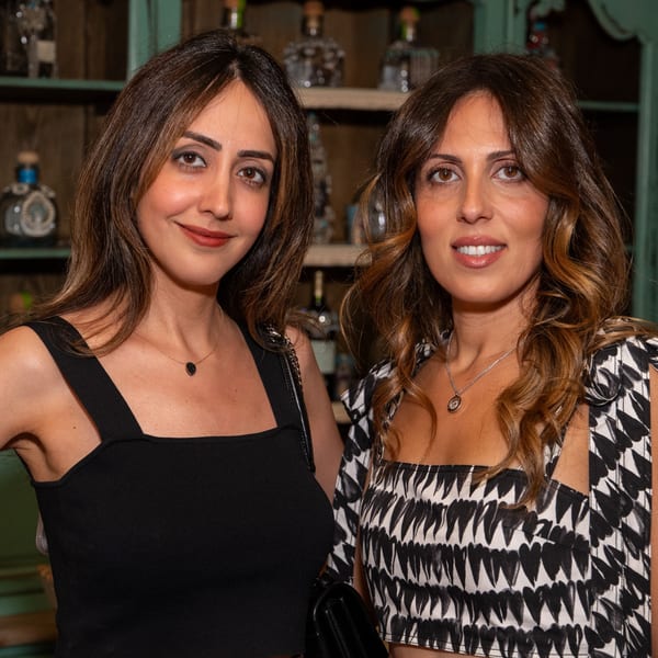 Two women with long brown hair smiling, one in a black top, the other in a patterned top.