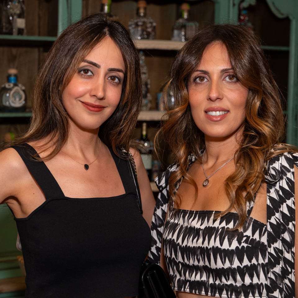 Two women with long brown hair smiling, one in a black top, the other in a patterned top.