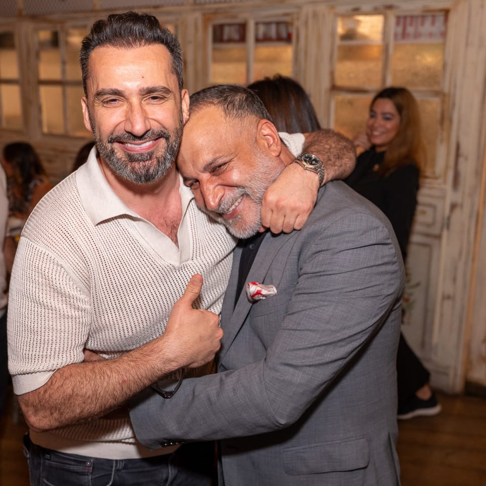Two smiling men embrace warmly, one in a white polo, the other in a gray suit, at Darvish Kitchen.