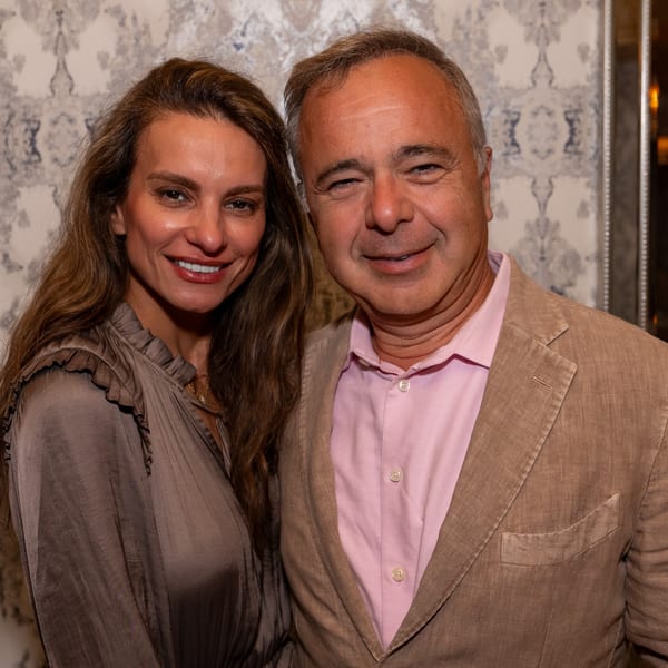A smiling man in a tan jacket and pink shirt with a woman in a brown ruffled top.