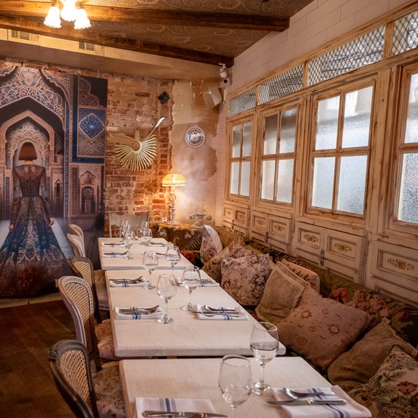Restaurant interior with set tables, patterned banquette seating, large windows, and a decorative wall mural.